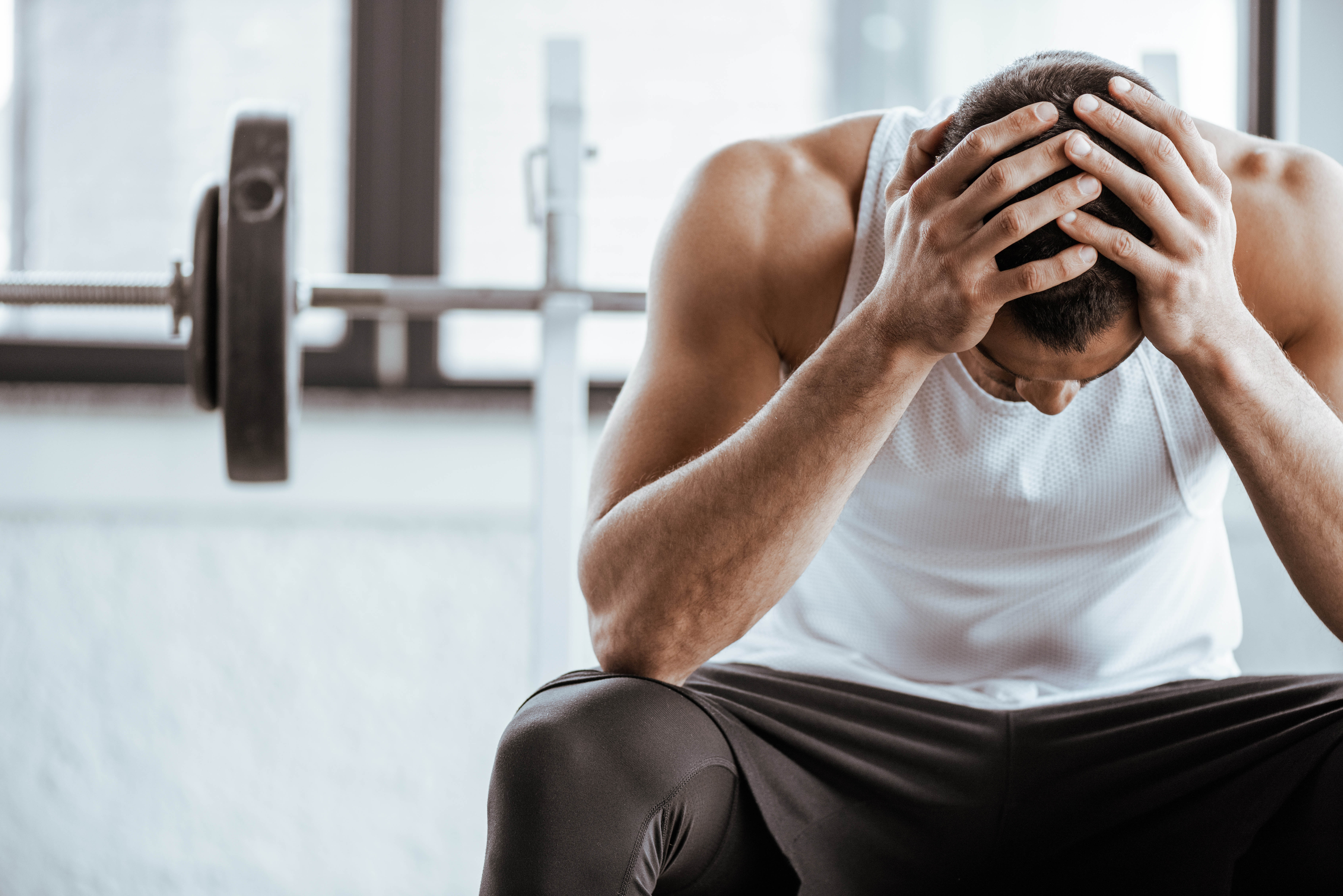Fit young man sitting in the gym with his head in the hands, weights in the background Fit young man sitting in the gym with his head in the hands, weights in the background